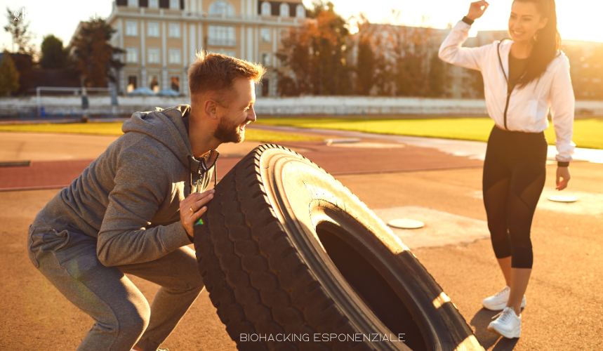 Allenamento outdoor con esercizio di forza e resistenza: uomo che solleva un pneumatico mentre una donna lo incoraggia, simbolo di energia, performance e biohacking.
